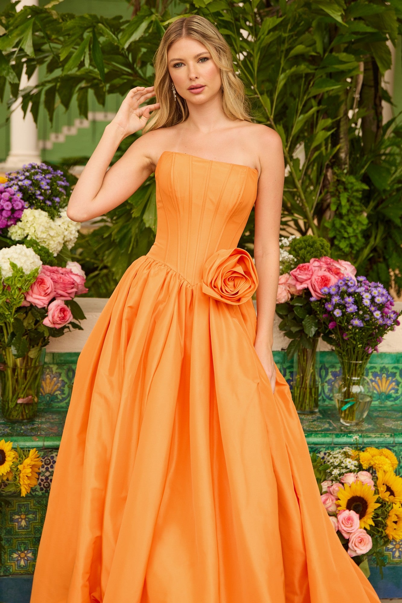 A woman in the Ava Presley 47204 long prom dress, featuring a strapless taffeta design with a corset bodice and large rose detail at the waist, stands before a lush floral background.